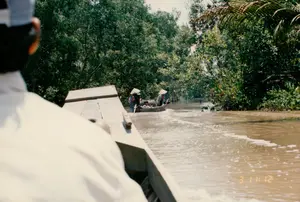 Trong ảnh có dòng chữ số màu vàng ở góc dưới bên phải: '3 11:12'.。Bối cảnh là một con kênh hoặc một con sông ở vùng nông thôn, có thể là ở Sóc Trăng. Nước sông có màu nâu phù sa. Hai bên bờ là thảm thực vật nhiệt đới rậm rạp, chủ yếu là cây xanh và có thể thấy cả những tàu lá dừa.。Hoạt động chính là di chuyển trên sông nước. Bức ảnh được chụp từ một chiếc ghe lớn, nhìn về phía trước. Ở phía xa hơn, có một chiếc xuồng gỗ nhỏ đang di chuyển trên sông. Trên chiếc xuồng này có một vài người, trong đó có ít nhất hai người lớn đang đội nón lá và một đứa trẻ. Một người đang chèo thuyền. Cảnh tượng này thể hiện cuộc sống sinh hoạt và giao thông đường thủy đặc trưng của người dân vùng Đồng bằng sông Cửu Long.。（日本語訳）。写真の右下の角に「3 11:12」という黄色い数字の列があります。背景は田舎の運河または川で、おそらくソクチャン省でしょう。川の水は沖積土で茶色です。両岸は鬱蒼とした熱帯植物で、主に緑の木々で、ヤシの葉も見られます。主な活動は水上での移動です。写真は大きなサンパン船から前方を向いて撮影されています。さらに遠くには、川の上を移動している小さな木製のボートがあります。このボートには数人が乗っており、その中にはノンラーをかぶった少なくとも2人の大人と1人の子供がいます。一人が船を漕いでいます。この光景は、メコンデルタ地域の人々の特徴的な日常生活と水上交通を表しています。