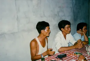 Bức ảnh chụp cảnh ba người đàn ông Việt Nam đang ngồi nói chuyện tại một chiếc bàn trong một căn phòng. Bức tường phía sau họ là tường xi măng thô, chưa được sơn. Trên tường có vài chữ viết mờ không thể đọc rõ. Người đàn ông ngồi ngoài cùng bên trái mặc một chiếc áo may ô (áo ba lỗ) màu trắng và đang nói chuyện, tay làm điệu bộ. Người đàn ông ngồi giữa mặc áo sơ mi trắng dài tay, và người đàn ông bên phải mặc áo sơ mi xanh nhạt cộc tay. Cả hai đang chăm chú lắng nghe. Trên bàn được trải một tấm khăn sọc caro đỏ và trắng. Trên bàn có một chiếc đồng hồ điện tử nhỏ hiển thị giờ là "2:34", một ấm trà bằng sứ, và một thiết bị điện tử nhỏ màu đen trông giống máy nhắn tin.。（日本語訳）。彼らの後ろの壁は、塗装されていない、むき出しのセメント壁です。壁には、はっきりと読めないいくつかの文字が書かれています。一番左に座っている男性は白いマイヨー（タンクトップ）を着ており、身振りを交えながら話しています。真ん中に座っている男性は白い長袖シャツを、右側の男性は水色の半袖シャツを着ています。二人とも熱心に耳を傾けています。テーブルには赤と白のチェック柄の布が敷かれています。テーブルの上には、「2:34」と表示された小さなデジタル時計、磁器の急須、そしてポケットベルのような小さな黒い電子機器があります。