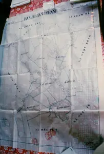 Bức ảnh chụp cận cảnh một tấm bản đồ hành chính cũ, có nhiều nếp gấp, được trải trên một chiếc bàn phủ khăn trải bàn hoa văn caro và hoa màu đỏ trắng. Đây là một bản đồ giấy, có thể được vẽ hoặc sao chép bằng tay, phản ánh công tác quản lý hành chính của địa phương vào khoảng năm 1994.。Các thông tin văn bản có thể đọc được trên bản đồ bao gồm:。- Tiêu đề lớn ở trên cùng: 'BẢN ĐỒ HÀNH CHÁNH'.。- Tên huyện: 'H. MỸ XUYÊN'.。- Tên các xã (Xã): 'XÃ THẠNH PHÚ', 'XÃ THAM ĐÔN', 'XÃ NGỌC ĐÔNG', 'XÃ NGỌC TỐ', 'XÃ GIA HÒA 1', 'XÃ HÒA TÚ 2'.。- Tên các ấp (Ấp): 'ẤP HÒA PHƯỚC', 'ẤP HÒA ĐI', 'ẤP HÒA NHÂN', 'ẤP HÒA TÂN', 'ẤP HÒA TRỰC', 'ẤP HÒA PHƯƠNG', 'ẤP HÒA TRUNG'.。- Ở góc dưới bên trái có một ô 'CHÚ THÍCH' nhưng nội dung không rõ.。Bản đồ mô tả ranh giới của các đơn vị hành chính cấp xã và ấp, cùng với một hệ thống sông ngòi và kênh rạch chằng chịt đặc trưng của vùng Đồng bằng sông Cửu Long. Các đường ranh giới và thủy văn được vẽ bằng mực xanh và đỏ. Việc tấm bản đồ được mở ra cho thấy nó đang được sử dụng để nghiên cứu, tham khảo hoặc thảo luận về các vấn đề liên quan đến địa lý và hành chính của khu vực.。（日本語訳）。赤と白のチェック柄と花柄のテーブルクロスが敷かれたテーブルの上に広げられた、多くの折り目がある古い行政地図のクローズアップ写真です。これは紙の地図で、手描きまたは手で複写された可能性があり、1994年頃の地方の行政管理業務を反映しています。地図上で読み取れるテキスト情報には、以下が含まれます。- 上部の大きなタイトル：「BẢN ĐỒ HÀNH CHÁNH」。- 県名：「H. MỸ XUYÊN」。- 各社（村）名（Xã）：「XÃ THẠNH PHÚ」、「XÃ THAM ĐÔN」、「XÃ NGỌC ĐÔNG」、「XÃ NGỌC TỐ」、「XÃ GIA HÒA 1」、「XÃ HÒA TÚ 2」。- 各邑（集落）名（Ấp）：「ẤP HÒA PHƯỚC」、「ẤP HÒA ĐI」、「ẤP HÒA NHÂN」、「ẤP HÒA TÂN」、「ẤP HÒA TRỰC」、「ẤP HÒA PHƯƠNG」、「ẤP HÒA TRUNG」。- 左下隅に「CHÚ THÍCH」の欄がありますが、内容は不明瞭です。この地図は、社（村）および邑（集落）レベルの行政単位の境界線と、メコンデルタ地域に特徴的な、複雑に張り巡らされた河川と運河のシステムを描写しています。境界線と水路は、青と赤のインクで描かれています。地図が広げられていることは、この地域の地理と行政に関連する問題を研究、参照、または議論するために使用されていることを示しています。