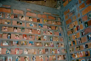 背景は、漆喰が塗られていない赤レンガの壁が二面ある、素朴に建てられた部屋の隅です。天井は波板トタンまたは同様の素材でできているようです。この二面の壁の表面には、小さなサイズのカラー写真がたくさん貼られています。これらの写真は、男性、女性、子供を含む様々な人々のポートレート写真で、様々な服装やポーズをとっています。いくつかの写真はセットされた背景のあるスタジオで撮影されていますが、他の写真は屋外で撮影されています。写真のファッションスタイルと色の質は、それらが1990年代頃に撮影されたことを示しています。このように多数の顧客のポートレート写真を展示していることは、ここが写真館またはカフェである可能性を示しており、装飾や自店のサービスの宣伝方法として写真を使用しています。画像の中には、読み取ることができる文字、テキスト、看板はありません