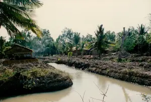 Bức ảnh chụp một khung cảnh nông thôn điển hình ở vùng sông nước, có thể là ở Sóc Trăng. Trung tâm của bức ảnh là một con kênh nhỏ (mương) với dòng nước màu nâu phù sa. Hai bên bờ kênh, đất được vun lên thành những bờ hay liếp vườn cao, cho thấy hoạt động cải tạo đất để canh tác nông nghiệp. Dọc theo bờ kênh là vài ngôi nhà lá đơn sơ, lợp bằng lá dừa, vách cũng làm từ vật liệu tự nhiên. Có một làn khói trắng bốc lên từ gần một ngôi nhà, có thể là từ việc nấu ăn hoặc đốt rác. Cảnh vật được bao phủ bởi cây cối xanh tươi, chủ yếu là những rặng dừa và các loại cây vườn khác. Không có bất kỳ biển hiệu hay chữ viết nào có thể nhận dạng được trong ảnh.。（日本語訳）。写真は、おそらくソクチャン省であろう、水郷地帯の典型的な田園風景を写しています。写真の中央には、沖積土で茶色く濁った水の小さな水路（用水路）があります。水路の両岸では、土が高く盛られて畝や畑の土手となっており、農業耕作のための土地改良活動が見て取れます。水路沿いには、屋根がヤシの葉で葺かれ、壁も自然素材でできた簡素な葉葺きの家が数軒あります。一軒の家の近くから白い煙が立ち上っており、炊事かゴミを燃やしている煙かもしれません。風景は青々とした木々に覆われており、主にヤシの木々やその他の園芸用の木々です。写真の中には、識別できる看板や文字は一切ありません。