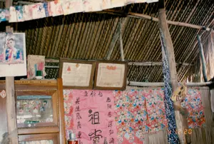 Bên trong một ngôi nhà truyền thống ở vùng nông thôn, có thể là ở Sóc Trăng. Ngôi nhà có kết cấu đơn sơ với mái lợp bằng lá và khung cột bằng gỗ. Cảnh chính trong ảnh là một bàn thờ tổ tiên. Một chiếc tủ thờ bằng gỗ có cửa kính, bên trong trưng bày nhiều ảnh nhỏ của gia đình. Trên nóc tủ có một cốc đựng nhang (hương) màu đỏ. Trên tường phía sau bàn thờ treo các vật phẩm trang trọng. Có hai bằng khen được đóng khung cẩn thận. Một bằng khen ghi rõ dòng chữ 'TỔ QUỐC GHI CÔNG', là giấy chứng nhận của nhà nước để ghi nhận công lao với đất nước. Bằng khen còn lại có chữ mờ, có thể đọc được một phần là '...CHƯƠNG KHÁNG CHIẾN'. Ở giữa là một tấm liễn thờ lớn màu hồng, trên đó viết chữ Hán. Chính giữa là hai chữ đại tự '祖 堂' (Tổ Đường). Hai bên là hai câu đối, bên trái là '義 生 成 義 重 山' (Nghĩa sinh thành nghĩa trọng sơn) và bên phải là '恩 鞠 恩 深 如 海' (Ân cúc ân sâu như hải), ca ngợi công ơn sinh thành và dưỡng dục của cha mẹ, tổ tiên. Ngoài ra, còn có các vật trang trí khác như một tấm lịch treo tường có hình một người phụ nữ, một dây treo các ảnh nhỏ gần mái nhà, và một phần của chiếc phích nước màu đỏ ở phía dưới.。（日本語訳）。ソクチャン省の田舎にある伝統的な家の中かもしれない。家は葉葺きの屋根と木製の柱でできた簡素な構造である。写真の主な光景は、先祖を祀る祭壇である。ガラス戸の付いた木製の祭壇用の棚があり、中には家族の小さな写真がたくさん飾られている。棚の上には、赤い色の線香立てが一つある。祭壇の後ろの壁には、格式ある品々が掛けられている。丁寧に額装された2枚の表彰状がある。一枚の表彰状には「TỔ QUỐC GHI CÔNG」（祖国は功績を記す）という文字がはっきりと書かれており、これは国への功労を認める国家の証明書である。もう一枚の表彰状は文字がかすれており、「...CHƯƠNG KHÁNG CHIẾN」（抗戦勲章）と一部読み取れる。中央には大きなピンク色の祭壇用の掛け軸があり、その上には漢字が書かれている。真ん中には「祖堂」（Tổ Đường）という二つの大きな文字がある。両側には二つの対句があり、左は「義生成義重山」（Nghĩa sinh thành nghĩa trọng sơn）、右は「恩鞠恩深如海」（Ân cúc ân sâu như hải）で、父母や先祖の生み育ててくれた恩を称えている。その他にも、女性の絵が描かれた壁掛けカレンダー、屋根の近くに吊るされた小さな写真の連なり、そして下の方には赤い魔法瓶の一部など、他の装飾品もある。