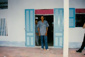 Trong ảnh có dấu thời gian kỹ thuật số là '17:52'. Bối cảnh là hiên trước của một ngôi nhà, có thể ở Sóc Trăng. Sàn hiên được lát bằng gạch tàu màu nâu đỏ. Có hai người đàn ông Việt Nam đang đứng ở cửa ra vào. Người đàn ông lớn tuổi hơn ở phía trước đang mỉm cười nhìn vào máy ảnh; ông mặc áo sơ mi cộc tay sọc ca rô xanh trắng, quần jean xanh và đi dép lê màu trắng. Phía sau ông, một người đàn ông khác đang đứng nép sau cánh cửa và nhìn ra. Cửa chính là loại cửa lá sách hai cánh, được sơn màu xanh lam và đang mở rộng. Phía trên khung cửa có treo một tấm rèm vải màu hồng. Bức tường của ngôi nhà được sơn màu trắng với phần chân tường màu xanh lam. Bên trái có một cửa sổ với các thanh chắn dọc.。（日本語訳）。写真には「17:52」というデジタルタイムスタンプがあります。背景は、おそらくソクチャン省にある家の玄関ポーチです。ポーチの床は赤褐色のテラコッタタイルで敷き詰められています。戸口に二人のベトナム人男性が立っています。手前にいる年配の男性はカメラを見て微笑んでいます。彼は青と白のチェック柄の半袖シャツ、青いジーンズを着用し、白いスリッパを履いています。彼の後ろでは、別の男性がドアの後ろに隠れるように立って外を見ています。正面のドアは両開きのルーバー扉で、青色に塗られており、大きく開いています。ドア枠の上には、ピンク色の布のカーテンが掛けられています。家の壁は白色に塗られており、巾木の部分は青色です。左側には縦格子付きの窓があります。