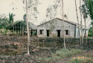 背景はソクチャン省のある農村地域で、一つの世帯かもしれません。写真の中央には、レンガまたはコンクリートの壁で建てられ、屋根は瓦葺きで、木製の鎧戸が付いた窓がある平屋があります。その隣には、木造の骨組みで、屋根はトタンまたは葉で葺かれた付属の建物があり、そこには衣類が干されています。家々の前には小さな水路または池があり、その岸は浚渫されたばかりの泥土で盛られています。風景は、土地または池の改良作業が行われていることを示しています。周囲には多くのヤシの木と、細い幹のユーカリの木が並んでいます。右奥には、一人の人が家の近くに立っています。写真の右下には「5 8:49」というタイムスタンプがあります