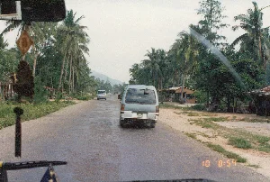 Bức ảnh được chụp từ bên trong một chiếc ô tô đang di chuyển trên một con đường ở vùng nông thôn Việt Nam vào năm 1996. Con đường nhựa hai làn, mặt đường còn ướt, cho thấy trời có thể vừa mưa xong. Hai bên đường rợp bóng những hàng dừa cao. Phía trước là một chiếc xe du lịch nhỏ (xe van) màu bạc. Ở phía xa hơn trên con đường, có thể thấy một chiếc xe van khác và một chiếc xe tải. Bên phải đường có một vài ngôi nhà hoặc quán nhỏ ven đường được xây dựng đơn sơ, có thể thấy vài người đang đứng ở đó. Phong cảnh phía xa là những dãy núi trập trùng. Góc dưới bên phải của ảnh có dấu thời gian kỹ thuật số màu cam ghi '10 8:54', cho biết thời điểm chụp là 8 giờ 54 phút ngày 10.。（日本語訳）。この写真は1996年にベトナムの田舎道で、移動中の車内から撮影されたものです。2車線のアスファルトの道路で、路面はまだ濡れており、雨が降ったばかりであることがうかがえます。道路の両側には、高いヤシの木が並び、影を落としています。前方には、銀色の小型乗用車（バン）が1台います。道路のさらに遠くには、別のバン1台とトラック1台が見えます。道路の右側には、簡素な作りの家や道端の小さな店がいくつかあり、数人がそこに立っているのが見えます。遠くの風景は、起伏のある山脈です。写真の右下隅には、「10 8:54」と記されたオレンジ色のデジタルタイムスタンプがあり、撮影時刻が10日の8時54分であることを示しています。