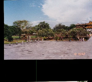 Bức ảnh chụp một khoảng sân rộng lớn được lát bằng đá, nằm trong một khu di tích lịch sử, có khả năng là Đại Nội Huế. Bầu trời trong xanh với những đám mây trắng. Phía sau sân là các công trình kiến trúc cổ kính với mái ngói men vàng đặc trưng của cung đình. Dọc theo rìa sân là hàng cây xanh, trong đó có nhiều cây hoa sứ (cây đại) đang nở hoa. Trên sân có các cột đá nhỏ được xếp ngay ngắn. Ở phía bên phải của bức ảnh, một nhóm nhỏ du khách đang đứng gần một công trình, trong đó có người đội nón lá. Dọc theo lối đi có cắm nhiều lá cờ. Dấu thời gian màu cam ở góc dưới bên phải của hình ảnh ghi '10 15:52'.。（日本語訳）。この写真は、歴史的遺跡の中にある、石で舗装された広大な中庭を写したもので、フエの王宮（ダイ・ノイ）である可能性が高い。空は青く澄み渡り、白い雲が浮かんでいる。中庭の奥には、宮廷特有の黄色い釉薬瓦の屋根を持つ古風な建築物がある。中庭の縁に沿って緑の木々が並んでおり、その中には花を咲かせているプルメリア（別名：cây đại）の木が多くある。中庭には、小さな石柱が整然と並べられている。写真の右側には、小さな観光客のグループが建物近くに立っており、その中にはノンラー（ベトナムの円錐形の帽子）をかぶっている人がいる。通路に沿って多くの旗が立てられている。画像の右下隅にあるオレンジ色のタイムスタンプには「10 15:52」と記されている。