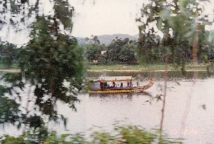 Bức ảnh chụp cảnh một con sông rộng, có khả năng là sông Hương ở Huế, vào khoảng năm 1996. Ở giữa sông là một chiếc thuyền rồng, một loại thuyền du lịch đặc trưng của khu vực này. Thuyền có màu vàng, mũi thuyền được chạm khắc hình đầu rồng, và có một khoang có mái che dành cho hành khách. Có vẻ như thuyền đang chở khách du lịch đi dạo trên sông. Bờ sông đối diện có cây cối xanh tốt, một vài ngôi nhà và những ngọn đồi ở phía xa. Bức ảnh được chụp qua những tán lá cây ở tiền cảnh, tạo ra hiệu ứng mờ do chuyển động. Ở góc dưới bên phải của hình ảnh có dấu thời gian '10 12:25'.。（日本語訳）。写真は、1996年頃、おそらくフエのフオン川と思われる、広い川の風景を撮影したものです。川の中央には、この地域特有の観光船である龍の船が一艘あります。船は黄色で、船首には龍の頭が彫られており、乗客用の屋根付きの船室があります。船は観光客を乗せて川を遊覧しているようです。対岸には緑豊かな木々、いくつかの家、そして遠くには丘が見えます。写真は前景の木の葉を通して撮影されており、動きによるブレ効果が生じています。画像の右下隅には「10 12:25」というタイムスタンプがあります。