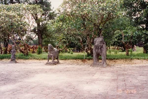 Bức ảnh chụp một không gian ngoài trời tại một khu di tích lịch sử, có khả năng là sân chầu trong một khu lăng tẩm ở Huế. Tiền cảnh là một khoảng sân rộng được lát bằng gạch đá vuông. Dọc theo rìa sân là một hàng tượng đá đặt trên bệ, bao gồm một tượng quan viên, một tượng ngựa và một tượng voi, tất cả đều được tạc một cách chi tiết. Đây là những bức tượng thường thấy trên đường thần đạo dẫn vào các lăng mộ vua chúa. Phía sau các bức tượng là một khu vườn với nhiều cây xanh, bao gồm cả cây thông và những cây hoa sứ (cây đại) đang nở hoa trắng rộ. Khung cảnh rất yên tĩnh và trang nghiêm, không có hoạt động nào của con người diễn ra. Ở góc dưới cùng bên phải của bức ảnh có một dấu thời gian kỹ thuật số màu cam hiển thị '10 12:46'.。（日本語訳）。この写真は、歴史的遺跡の屋外空間を撮影したもので、フエの陵墓群にある拝謁の庭である可能性があります。前景には、四角い石のタイルで舗装された広い庭があります。庭の端に沿って、台座の上に置かれた石像の列があり、文官の像、馬の像、象の像が含まれ、すべてが精巧に彫られています。これらは、皇帝の陵墓へと続く神道（参道）でよく見られる像です。像の後ろには、松の木や、白い花を咲かせているプルメリア（cây đại）の木など、多くの緑の木々がある庭園が広がっています。風景は非常に静かで荘厳であり、人間の活動は見られません。写真の右下隅には、「10 12:46」と表示されたオレンジ色のデジタルタイムスタンプがあります。