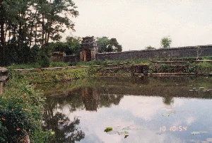 Trên ảnh có dấu thời gian kỹ thuật số màu cam "10 10:54".。Đây là khung cảnh tại một di tích lịch sử, có khả năng là một khu lăng tẩm ở Huế. Ở phía trước là một hồ nước lớn, mặt nước phẳng lặng, trên mặt hồ có những chiếc lá súng. Mặt nước phản chiếu rõ nét hình ảnh của bầu trời, cây cối và một chiếc cổng thành cổ ở phía xa. Bao quanh hồ là bờ kè bằng đá và đất, cỏ dại mọc um tùm. Ở hậu cảnh là một bức tường thành cổ, dài và rêu phong. Nổi bật trên bức tường là một chiếc cổng thành cổ kính với kiến trúc truyền thống, có nhiều tầng mái cong. Phía sau tường thành là những hàng cây thông và cây cối rậm rạp. Toàn bộ khung cảnh toát lên vẻ yên tĩnh, cổ kính. Không có hoạt động nào của con người được ghi nhận trong ảnh.。（日本語訳）。写真にはオレンジ色のデジタルタイムスタンプ「10 10:54」があります。これは史跡の光景で、フエの陵墓群である可能性があります。手前には大きな池があり、水面は穏やかで、水面にはスイレンの葉が浮かんでいます。水面には、空、木々、そして遠くにある古い城門の姿がはっきりと映り込んでいます。池の周りは石と土でできた土手で、雑草が生い茂っています。背景には、長く苔むした古い城壁があります。城壁の上で際立っているのは、伝統的な建築様式で、幾層にもなった湾曲した屋根を持つ古風な城門です。城壁の後ろには、松の木々や鬱蒼とした木々が並んでいます。全体の風景は、静かで古風な趣を醸し出しています。写真には人間の活動は記録されていません。