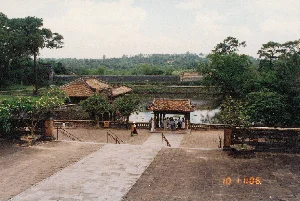 Bức ảnh chụp một khung cảnh tại một khu di tích lịch sử, có khả năng là một trong những lăng tẩm của vua triều Nguyễn ở Huế, được chụp vào năm 1996. Quang cảnh được chụp từ trên cao, nhìn xuống một khoảng sân rộng lát gạch và một hồ nước.。Hoạt động chính là tham quan du lịch. Một nhóm du khách đang tụ tập tại một nhà thủy tạ (phương đình) bằng gỗ, có mái ngói, nằm ngay cạnh bờ hồ. Trong nhóm có một người phụ nữ mặc áo dài trắng. Một vài người khác đang đi dạo trong sân.。Các vật thể nổi bật bao gồm các công trình kiến trúc cổ với mái ngói âm dương, nhà thủy tạ, hồ nước tĩnh lặng, và những bức tường thành bao quanh. Cảnh quan thiên nhiên có nhiều cây xanh tươi tốt như cây thông và cây hoa sứ (cây đại) đang nở hoa.。Về chữ viết, ở góc dưới bên phải hình ảnh có dấu thời gian kỹ thuật số màu cam ghi '10 11:06'. Không có bảng hiệu, bia đá hay chữ viết nào khác có thể đọc được.。（日本語訳）。この写真は、1996年に撮影された、フエにあるグエン朝の皇帝陵の一つである可能性が高い、ある史跡の風景を写したものです。風景は上から撮影されており、レンガ敷きの広い中庭と池を見下ろしています。主な活動は観光です。瓦屋根を持つ木造の水上家屋（方亭）に観光客のグループが集まっています。その建物は池のほとりにあります。グループの中には、白いアオザイを着た女性が一人います。他の数人が中庭を散策しています。目立つ物としては、陰陽瓦の屋根を持つ古い建築物、水上家屋、静かな池、そして周囲を囲む城壁などがあります。自然景観には、松や花を咲かせているプルメリア（大木）のような青々とした多くの木々があります。文字については、画像の右下隅にオレンジ色のデジタルタイムスタンプで「'10 11:06」と記されています。他に読み取れる看板、石碑、文字はありません。