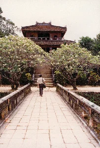 Bức ảnh chụp một khung cảnh tại một di tích lịch sử, có thể là một lăng tẩm hoặc một phần của Đại Nội Huế. Một người đàn ông đang đi bộ trên một cây cầu đá, tiến về phía một ngôi lầu cổ hai tầng. Ngôi lầu này có kiến trúc cung đình đặc trưng với mái ngói âm dương và các chi tiết trang trí bằng gỗ. Dẫn lên ngôi lầu là một cầu thang bằng đá. Hai bên lối đi là hai cây hoa sứ (hoa đại) lớn đang nở rộ hoa trắng. Cây cầu cũng được xây dựng cổ kính với lan can được trang trí. Không có văn bản nào có thể đọc được trong ảnh.。（日本語訳）。写真は、歴史的遺跡、おそらくは帝廟かフエの王宮の一部で撮影された風景を写しています。一人の男性が石橋の上を歩いており、二階建ての古い楼閣に向かって進んでいます。この楼閣は、陰陽瓦の屋根と木製の装飾的な細部を持つ、特徴的な宮廷建築様式です。楼閣へは石の階段が続いています。通路の両側には、白い花を咲かせている2本の大きなプルメリア（hoa sứ/hoa đại）の木があります。橋もまた古風に建てられており、手すりには装飾が施されています。写真の中には読み取れる文字はありません。