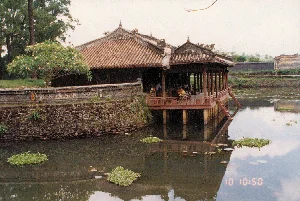 Bối cảnh là một công trình kiến trúc cổ trong một khu di tích lịch sử, có khả năng là ở Huế, dựa trên phong cách kiến trúc. Hình ảnh cho thấy một nhà thủy tạ (nhà tạ) bằng gỗ với mái ngói âm dương màu nâu đỏ, được xây dựng nhô ra trên một hồ nước hoặc hào nước. Kiến trúc này mang đậm nét cung đình triều Nguyễn. Bên trong nhà tạ, có một vài người đang ngồi thư giãn. Mặt hồ tĩnh lặng, có những cụm bèo tây (lục bình) và lá súng trôi nổi, phản chiếu bóng của công trình và bầu trời. Bờ hồ được xây bằng đá và gạch. Trên bờ có cây cối, bao gồm một cây hoa sứ đang nở hoa. Không có văn bản nào có thể đọc được ngoại trừ dấu thời gian kỹ thuật số "10 10:50" của máy ảnh được in trên mặt nước.。（日本語訳）。背景は歴史遺跡地区にある古い建築物で、建築様式からフエにある可能性があります。写真には、赤褐色の陰陽瓦の屋根を持つ木造の水亭（涼亭）が写っており、湖または堀の上に突き出るように建てられています。この建築はグエン朝の宮廷様式を色濃く反映しています。亭の中では、数人が座ってくつろいでいます。湖面は静かで、ホテイアオイ（ウォーターヒヤシンス）の群生とスイレンの葉が浮かんでおり、建物と空の影を映しています。湖の岸は石とレンガで造られています。岸には木々があり、その中には花を咲かせているプルメリアの木も含まれています。水面に印字されたカメラのデジタルタイムスタンプ「10 10:50」を除いて、読み取れる文字はありません。