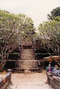 Bức ảnh chụp một lối vào một công trình kiến trúc cổ, có thể là một ngôi chùa hoặc lăng tẩm ở khu vực Huế. Chính giữa là một cầu thang bằng đá rộng, dẫn lên một cổng tam quan cổ kính có mái ngói. Hai bên lối đi là hai cây hoa sứ (cây đại) lớn đang nở hoa trắng. Bên phải, dưới gốc cây, có một quầy hàng nhỏ bán nước giải khát. Một người phụ nữ đội mũ đang đứng bán hàng bên cạnh chiếc bàn có trải khăn caro xanh trắng, trên bàn bày các loại chai nước ngọt. Bên cạnh có một thùng đá và vài chiếc ghế nhựa. Một chiếc ô lớn dùng để che nắng cho quầy hàng. Ở phía trước có lan can của một cây cầu nhỏ được trang trí công phu. Trên các cột của cổng chính có thể thấy các ký tự chữ Hán hoặc Nôm nhưng không thể nhận dạng rõ.。（日本語訳）。写真は、フエ地方にある寺院か廟と思われる古い建築物への入り口を写しています。中央には幅の広い石の階段があり、瓦屋根を持つ古風な三関門へと続いています。通路の両側には、白い花を咲かせている2本の大きなプルメリア（インドソケイ）の木があります。右側、木の根元には、清涼飲料水を売る小さな売店があります。帽子をかぶった女性が、青と白のチェック柄のテーブルクロスが敷かれたテーブルの横で商品を売っており、テーブルの上には様々な種類のペットボトル飲料が並べられています。その隣には、クーラーボックスといくつかのプラスチック製の椅子があります。大きな傘が売店の日よけに使われています。手前には、精巧に装飾された小さな橋の欄干があります。正門の柱には漢字かチュノムの文字が見えますが、はっきりと識別することはできません。