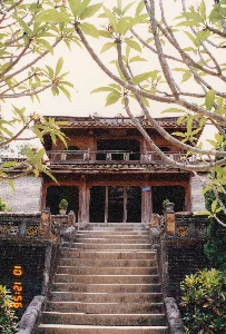 Bức ảnh chụp một công trình kiến trúc cổ hai tầng, có khả năng là một ngôi nhà hoặc một phần của khu lăng tẩm, di tích lịch sử ở Huế. Một cầu thang đá rộng dẫn lên tòa nhà. Toàn bộ công trình có kết cấu bằng gỗ và gạch, với mái ngói âm dương đặc trưng của kiến trúc truyền thống Việt Nam. Tầng một là không gian mở với các cột gỗ lớn. Tầng hai có ban công với lan can được trang trí. Hai bên cầu thang là lan can bằng đá được chạm khắc tinh xảo, và trên các bức tường thấp có các họa tiết trang trí, bao gồm cả biểu tượng chữ Thọ. Ở chiếu nghỉ trên cùng của cầu thang có đặt hai chậu cây cảnh. Có một tấm biển nhỏ màu xanh gắn trên cột gỗ nhưng không đọc được chữ. Phía trên ban công tầng hai cũng có một bức hoành phi nhưng chữ viết trên đó không thể nhận dạng. Ở phía trước, các cành của một cây hoa sứ (cây hoa đại) đang nở hoa tạo thành một khung cảnh tự nhiên cho tòa nhà. Không có người xuất hiện trong ảnh, khung cảnh mang vẻ tĩnh lặng, cổ kính. Ở góc dưới bên trái của bức ảnh có dấu thời gian kỹ thuật số màu cam ghi "10 12:56 '96".。（日本語訳）。写真は2階建ての古い建築物を写しており、フエの家屋または陵墓群、歴史的遺跡の一部である可能性がある。幅の広い石の階段が建物へと続いている。建物全体は木とレンガで構成され、ベトナムの伝統建築に特徴的な陰陽瓦の屋根を持つ。1階は大きな木の柱がある開放的な空間である。2階には装飾が施された手すりのあるバルコニーがある。階段の両側には精巧に彫刻された石の手すりがあり、低い壁には「寿」の字のシンボルを含む装飾模様が施されている。木の柱には小さな青い看板が取り付けられているが、文字は読み取れない。2階のバルコニーの上にも扁額があるが、そこに書かれている文字は判別できない。手前では、花を咲かせたプルメリアの木の枝が、建物の自然な額縁を形成している。写真には人物は写っておらず、景色は静かで古風な趣を醸し出している。写真の左下隅には、「10 12:56 '96」と記されたオレンジ色のデジタルタイムスタンプがある。