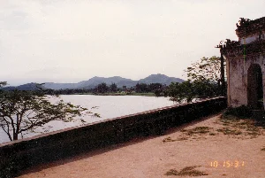 Bức ảnh chụp một khung cảnh thiên nhiên và kiến trúc cổ, có khả năng là tại một khu di tích lịch sử ở Huế, vào năm 1996.。Về chữ viết, ở góc dưới bên phải có dấu thời gian kỹ thuật số của máy ảnh là "10 15:31". Không có chữ viết nào khác trên các công trình hay biển báo.。Về cảnh quan, đây là một không gian yên tĩnh nhìn ra một hồ nước lớn và phẳng lặng. Phía xa là những dãy núi nhấp nhô dưới bầu trời có mây. Bên phải của bức ảnh là một phần của một công trình kiến trúc cổ, có thể là một cổng tam quan hoặc một vọng lâu, với những bức tường đã cũ và mái ngói cong theo phong cách truyền thống. Một bức tường lan can thấp chạy dọc theo bờ hồ, ngăn cách với một khoảng sân đất rộng. Cây cối, bao gồm một cây lớn với nhiều cành ở phía trước, tạo thêm vẻ thanh bình cho cảnh vật. Không có người hay hoạt động nào được ghi lại, chỉ có một phong cảnh tĩnh lặng và trầm mặc.。（日本語訳）。この写真は、1996年にフエの歴史遺跡である可能性のある場所で撮影された、自然と古い建築物の風景を写したものです。文字については、右下隅にカメラのデジタルタイムスタンプ「10 15:31」があります。建造物や看板には他に文字はありません。景観については、これは大きく穏やかな湖を見渡す静かな空間です。遠くには、雲のある空の下に起伏のある山々が連なっています。写真の右側には、古い建築物の一部があり、それは三関門（タムクアン門）か望楼かもしれません。古びた壁と伝統的な様式の湾曲した瓦屋根を持っています。低い欄干の壁が湖岸に沿って伸びており、広々とした土の庭とを隔てています。前方にある多くの枝を持つ一本の大木を含む木々が、風景にさらなる静けさを加えています。人や活動は記録されておらず、ただ静寂で物静かな風景があるだけです。