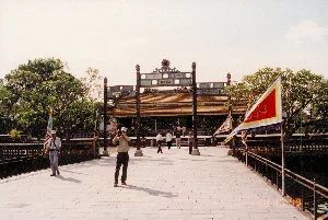 Bức ảnh chụp tại một khu di tích lịch sử ở Huế, có khả năng là một trong các lăng tẩm triều Nguyễn. Khung cảnh cho thấy một cây cầu đá rộng bắc qua một hồ nước, dẫn đến một công trình kiến trúc cổ. Trên cầu có một số du khách đang tham quan. Một người đàn ông ở tiền cảnh đang cầm máy ảnh để chụp hình. Một du khách khác đội mũ trắng đang đi bộ trên cầu. Ở phía xa, nhiều người đang đi bộ về phía cổng. Dọc hai bên thành cầu cắm nhiều lá cờ lễ hội truyền thống nhiều màu sắc (cờ ngũ sắc), trên một số lá cờ có họa tiết hình rồng. Phía cuối cây cầu là một cổng tam quan uy nghi với mái ngói. Trên tấm biển ở giữa cổng có khắc bốn chữ Hán là 「明光大正」 (Minh Quang Đại Chính). Cây cối xanh tươi bao quanh khu di tích.。（日本語訳）。写真はフエの歴史遺跡で撮影されたもので、おそらくグエン朝の陵墓の一つでしょう。風景には、湖を渡る広い石橋が写っており、古い建築物へと続いています。橋の上には数人の観光客が観光しています。前景には、男性がカメラを持って写真を撮っています。別の白い帽子をかぶった観光客が橋の上を歩いています。遠くでは、多くの人々が門に向かって歩いています。橋の欄干の両側には、色とりどりの伝統的な祭りの旗（五色旗）が数多く立てられており、いくつかの旗には龍の模様が描かれています。橋の先には、瓦屋根の荘厳な三関門があります。門の中央にある扁額には、「明光大正」（ミン・クアン・ダイ・チン）という四つの漢字が刻まれています。青々とした木々が史跡を囲んでいます。