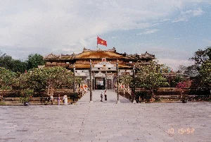 Bức ảnh chụp quang cảnh Ngọ Môn, cổng chính của Hoàng thành Huế, vào một buổi chiều. Trên đỉnh cổng chính có treo lá quốc kỳ Việt Nam màu đỏ sao vàng. Trên trán cổng có khắc hai chữ Hán lớn là "午門" (Ngọ Môn).。Đây là một địa điểm di tích lịch sử, với một công trình kiến trúc cổ có mái ngói màu vàng đặc trưng. Phía trước cổng là một sân rộng lát đá. Một lối đi dẫn vào cổng được trang trí bằng nhiều lá cờ hội nhiều màu sắc cắm dọc hai bên. Một vài nhóm người, có lẽ là du khách, đang có mặt tại đây. Ở phía bên trái, có một nhóm người đang đứng, trong đó có phụ nữ mặc áo dài và đội nón lá. Ở trung tâm, hai người đàn ông đang đi bộ về phía cổng. Xung quanh sân có trồng nhiều cây xanh, bao gồm cả những cây hoa sứ (hoa đại) đang nở hoa. Có thể nhìn thấy một chiếc xe khách hoặc xe buýt qua lối vào của cổng.。（日本語訳）。写真は、ある日の午後、フエ王宮の正門である午門（ゴ・モン）の光景を撮影したものです。正門の上には、金星紅旗（黄色の星の赤いベトナム国旗）が掲げられています。門の額には「午門」（ゴ・モン）という二つの大きな漢字が刻まれています。これは歴史的な遺跡であり、特徴的な黄色の瓦屋根を持つ古い建築物です。門の前には石畳の広い広場があります。門へと続く通路は、両脇に立てられた色とりどりの多くの祭りの旗で飾られています。おそらく観光客と思われるいくつかのグループがここにいます。左側には、立っている人々のグループがおり、その中にはアオザイを着用しノンラーをかぶった女性たちがいます。中央では、二人の男性が門に向かって歩いています。広場の周りには多くの緑の木が植えられており、その中には花を咲かせているプルメリア（ホア・ダイ）の木も含まれています。門の入り口を通して、観光バスまたは路線バスが見えます。