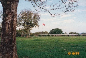 Bức ảnh chụp một không gian rộng lớn với bãi cỏ xanh mướt, có khả năng là khu vực phía trước Kinh thành Huế. Ở phía trước bên trái là một thân cây cổ thụ to lớn. Phía xa là hình ảnh Cột cờ Huế (Kỳ Đài) sừng sững trên một phần của tường thành. Lá cờ đỏ sao vàng của Việt Nam đang tung bay trên đỉnh cột cờ. Bầu trời trong xanh, có vài gợn mây. Khung cảnh rất yên tĩnh và trang nghiêm, không có hoạt động nào của con người diễn ra. Ở góc dưới bên phải bức ảnh có dấu thời gian màu vàng hiển thị '10 16:29'.。（日本語訳）。写真は、緑豊かな芝生のある広大な空間を写しており、フエの王宮前のエリアである可能性があります。左手前には、大きな古い木の幹があります。遠方には、城壁の一部の上に堂々とそびえ立つフエの旗竿（午門前の旗台）の姿があります。ベトナムの金星紅旗が旗竿の頂上で翻っています。空は青く澄み渡り、いくつかの筋雲があります。風景は非常に静かで荘厳であり、人間の活動は見られません。写真の右下隅には、「10 16:29」と表示された黄色のタイムスタンプがあります。