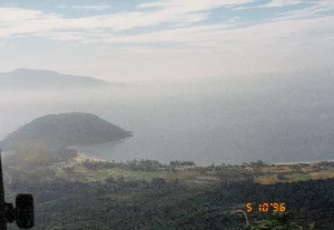 Bức ảnh chụp toàn cảnh một vùng ven biển nhìn từ trên cao, có khả năng được chụp từ đèo Hải Vân nhìn về phía vịnh Lăng Cô hoặc Đà Nẵng. Phong cảnh bao gồm một vịnh biển, một bán đảo nhỏ được cây cối bao phủ, và một dải bờ biển có bãi cát. Ở khu vực đồng bằng ven biển, có thể thấy rõ những thửa ruộng nông nghiệp được chia ô, cho thấy hoạt động canh tác của người dân. Phía xa, những dãy núi chìm trong một lớp sương mù mỏng. Bầu trời có mây. Ở góc dưới bên phải của bức ảnh có một dấu ngày kỹ thuật số màu cam ghi '5 10'96', cho biết bức ảnh được chụp vào ngày 10 tháng 5 năm 1996. Khung cảnh mang vẻ đẹp hoang sơ, yên bình của thiên nhiên Việt Nam.。（日本語訳）。高所から見た沿岸地域の一帯を写したパノラマ写真で、ハイヴァン峠からランコー湾またはダナンの方角を望んで撮影された可能性があります。風景には、湾、木々に覆われた小さな半島、そして砂浜のある海岸線が含まれています。沿岸の平野部では、区画に分けられた農地がはっきりと見え、人々の耕作活動を示しています。遠くには、山脈が薄い霧の中に沈んでいます。空には雲があります。写真の右下隅には、「5 10'96」と記されたオレンジ色のデジタル日付スタンプがあり、写真が1996年5月10日に撮影されたことを示しています。その光景は、ベトナムの自然が持つ、手付かずで穏やかな美しさを帯びています。