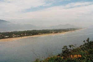 Trên ảnh có dấu ngày tháng màu cam "5 10 '96", cho biết ảnh được chụp vào ngày 10 tháng 5 năm 1996.。Bức ảnh ghi lại một khung cảnh ven biển tuyệt đẹp, được chụp từ một vị trí trên cao, có khả năng là từ một con đèo như Đèo Hải Vân, nhìn xuống một vịnh hoặc đầm phá. Cảnh vật chính là một dải đất hẹp và dài, một bên là biển cả mênh mông với sóng nhẹ, một bên là vùng đầm phá nước lặng. Dải đất này có một bãi biển cát vàng.。Một làng chài nhỏ nằm dọc theo dải đất, với những ngôi nhà thấp tầng nằm ẩn mình dưới những rặng cây xanh tốt. Có thể thấy một vài chiếc thuyền (ghe) nhỏ của ngư dân đang neo đậu trên bãi cát và trên mặt nước của đầm phá. Sự hiện diện của làng chài và thuyền bè cho thấy hoạt động chính của người dân nơi đây là đánh bắt thủy sản.。Ở phía xa, những dãy núi trập trùng hiện ra mờ ảo trong làn sương. Bầu trời có mây, tạo nên một không gian rộng lớn và yên bình, đặc trưng cho vùng duyên hải miền Trung Việt Nam. Ở tiền cảnh, góc dưới bên phải, là những bụi cây và hoa dại đang nở.。（日本語訳）。写真にはオレンジ色の「5 10 '96」という日付スタンプがあり、写真が1996年5月10日に撮影されたことを示しています。この写真は、ハイヴァン峠のような高所から湾またはラグーンを見下ろして撮影された、美しい海岸の風景を捉えています。主な風景は、片側が穏やかな波の広大な海、もう片側が静かな水のラグーン地帯となっている、細長い土地です。この土地には黄色い砂浜があります。土地に沿って小さな漁村があり、青々とした木々の下に低層の家々が隠れています。砂浜やラグーンの水面に停泊している漁師の小さな船（ボート）がいくつか見えます。漁村と船の存在は、ここの人々の主な活動が水産物の漁獲であることを示しています。遠くには、霧の中にぼんやりと連なる山々が現れています。空は曇っており、ベトナム中部の沿岸地域に特有の、広大で穏やかな空間を作り出しています。前景の右下隅には、茂みと咲いている野花があります。