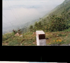 Chữ viết trên ảnh: '5 10 '96'.。Bối cảnh là một con đường đèo núi, có khả năng là Đèo Hải Vân, nhìn từ trên cao xuống thung lũng. Bức ảnh dường như được chụp từ một phương tiện đang di chuyển, vì phần tiền cảnh bị nhòe. Bên phải là sườn núi dốc được bao phủ bởi cây cối xanh tươi. Phía dưới thung lũng và ở phía xa, những dãy núi trập trùng được bao phủ bởi một lớp sương mù hoặc mây dày đặc. Có thể nhìn thấy một cọc tiêu bê tông ở ven đường trong tiền cảnh. Phía trên bên trái có các đường dây điện bắc qua. Phong cảnh mang vẻ hùng vĩ và mờ ảo đặc trưng của vùng núi.。（日本語訳）。写真上の文字：「'96年10月5日」。背景は山道の峠で、ハイヴァン峠の可能性があり、高所から谷を見下ろしている。写真は移動中の乗り物から撮影されたようで、前景がぼやけている。右側は、青々とした木々に覆われた急な山の斜面である。谷の下方と遠方には、幾重にも連なる山々が濃い霧または雲の層に覆われている。左上には電線が横切っている。風景は、山岳地帯特有の雄大で幻想的な趣を帯びている。