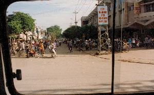 Bức ảnh được chụp từ bên trong một phương tiện giao thông, nhìn ra một con đường phố đông đúc ở Việt Nam vào năm 1996. Trên đường có rất nhiều người đang di chuyển bằng xe đạp và một số ít xe máy. Nhiều người, bao gồm cả phụ nữ đội nón lá, đang đi xe đạp với những chiếc giỏ lớn phía sau, cho thấy họ có thể đang đi chợ hoặc vận chuyển hàng hóa. Khung cảnh hai bên đường là những tòa nhà nhiều tầng, có các cửa hàng ở tầng trệt. Có một tấm biển hiệu lớn màu trắng với dòng chữ đỏ mang tính cổ động: "SỐNG VÀ LÀM VIỆC THEO HIẾN PHÁP VÀ PHÁP LUẬT". Trên một tòa nhà khác, có một biển quảng cáo màu vàng và đỏ cho sản phẩm "MÌ ĂN LIỀN LUCKY AN THAI" và một biển hiệu cửa hàng ghi "KIỀU OANH - HẠNH". Ngoài ra còn có một biển báo giao thông hình tam giác cảnh báo người đi bộ. Hệ thống cột điện và dây điện chằng chịt phía trên đường phố là một hình ảnh đặc trưng.。（日本語訳）。この写真は、1996年にベトナムの混雑した通りを、ある乗り物の中から撮影したものです。通りには、自転車で移動している多くの人々と、少数のバイクがいます。ノンラーをかぶった女性を含む多くの人々が、後ろに大きなかごを付けた自転車に乗っており、市場へ行く途中か、商品を運んでいる途中であることがうかがえます。通りの両側の風景は、1階に店舗が入った多層階の建物です。「憲法と法律に従って生き、働く」というスローガン的な赤い文字が書かれた大きな白い看板があります。別の建物には、「MÌ ĂN LIỀN LUCKY AN THAI」（ラッキー・アンタイ・インスタントラーメン）という商品の黄色と赤の広告看板と、「KIỀU OANH - HẠNH」と書かれた店の看板があります。その他に、歩行者に注意を促す三角形の交通標識もあります。通りの上空に張り巡らされた電柱と電線のシステムは、特徴的な光景です。