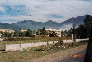 Bức ảnh chụp một khung cảnh ven đường với dãy núi hùng vĩ làm nền, có thể là ở khu vực gần đèo Hải Vân. Ở trung tâm là một tòa nhà nhiều tầng màu vàng, trên nóc có biển hiệu chữ đỏ ghi "CÔNG TY XI MĂNG HẢI VÂN". Xung quanh tòa nhà có cây cối, bao gồm cả cây dừa. Có một cột khói hoặc sương mù bốc lên từ phía sau tòa nhà hoặc từ sườn núi. Tiền cảnh là một con đường đất chạy dọc theo một bức tường bê tông thấp. Phía sau bức tường là một khu đất trống có cỏ mọc. Bầu trời trong xanh có mây và có nhiều đường dây điện chạy ngang qua. Ở góc dưới bên phải của ảnh có dấu ngày tháng kỹ thuật số màu cam "5 10 '96", cho thấy bức ảnh được chụp vào ngày 10 tháng 5 năm 1996.。（日本語訳）。写真は、雄大な山脈を背景にした道端の風景を写しており、おそらくハイヴァン峠付近の地域で撮影されたものでしょう。中央には黄色い多層階の建物があり、その屋上には「CÔNG TY XI MĂNG HẢI VÂN」と書かれた赤い文字の看板があります。建物の周りには、ヤシの木を含む木々があります。建物の後ろか山腹から、煙か霧の柱が立ち上っています。前景には、低いコンクリートの壁に沿って走る未舗装の道があります。壁の後ろには、草が生えている空き地があります。青空には雲があり、多くの電線が横切っています。写真の右下隅にはオレンジ色のデジタル日付スタンプ「5 10 '96」があり、これは写真が1996年5月10日に撮影されたことを示しています。