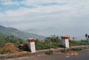 Trên ảnh có dấu ngày tháng màu cam '5 10 '96', cho thấy ảnh được chụp vào ngày 10 tháng 5 năm 1996. Đây là khung cảnh nhìn từ một con đường đèo, rất có thể là Đèo Hải Vân trên tuyến đường giữa Huế và Đà Nẵng. Ở phía trước là lề đường có hai cọc tiêu bê tông sơn trắng và đỏ, một loại cọc tiêu chỉ giới đường bộ phổ biến ở Việt Nam. Ven đường có các bụi cây dại mọc trên nền đất. Nhìn ra xa là một khung cảnh núi non trùng điệp, bao phủ bởi cây cối xanh tươi. Phía xa hơn, dưới thung lũng, có thể thấy một vùng vịnh hoặc bờ biển trải dài, với những dãy núi mờ ảo trong sương. Một con đường hoặc đường ray xe lửa dường như chạy dọc theo bờ biển. Bầu trời xanh, có mây trắng. Ở bên trái có vài đường dây điện chạy qua. Không có hoạt động nào của con người được ghi lại, chỉ có cảnh quan thiên nhiên hùng vĩ.。（日本語訳）。写真にはオレンジ色の「'96 5 10」という日付スタンプがあり、写真が1996年5月10日に撮影されたことを示しています。これは峠道から見た風景で、フエとダナンを結ぶルート上にあるハイヴァン峠である可能性が非常に高いです。手前には路肩があり、白と赤に塗られたコンクリート製のデリニエーターが2本あります。これはベトナムで一般的な道路境界を示す杭の一種です。道端には土の上に雑草の茂みが生えています。遠くを見ると、青々とした木々に覆われた山々が連なる風景が広がっています。さらに遠く、谷の下には、霧の中にぼんやりと浮かぶ山脈を伴う、広がる湾または海岸線が見えます。一本の道または線路が海岸線に沿って走っているように見えます。空は青く、白い雲があります。左側には数本の電線が通っています。人間の活動は記録されておらず、雄大な自然の景観だけがあります。