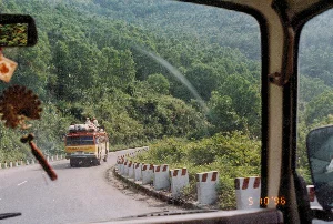 Trên ảnh có dấu ngày tháng kỹ thuật số màu cam ở góc dưới bên phải: '5 10 '96'. Phía sau chiếc xe khách có chữ và số nhưng khó đọc, có thể thấy '43A'.。Bức ảnh được chụp từ bên trong một chiếc ô tô đang di chuyển trên một con đường đèo núi quanh co, có khả năng là Đèo Hải Vân. Khung cảnh xung quanh là những ngọn đồi núi được bao phủ bởi rừng cây xanh rậm rạp.。Hoạt động chính là cảnh giao thông vận tải trên đèo. Một chiếc xe khách (xe đò) kiểu cũ, sơn màu vàng và đỏ, đang chạy phía trước. Trên nóc xe khách chất đầy hành lý, bao tải và hàng hóa. Đặc biệt, có một vài hành khách đang ngồi trên nóc xe cùng với đống hàng hóa, một cảnh tượng phổ biến trong giao thông ở Việt Nam vào thời điểm đó. Dọc theo con đường là một hàng cọc tiêu bê tông sơn sọc trắng đỏ. Từ góc nhìn của người chụp, có thể thấy một phần nội thất của chiếc xe, bao gồm gương chiếu hậu và một vật trang trí có tua rua đang treo.。（日本語訳）。写真の右下隅には、オレンジ色のデジタル日付スタンプ「'96 10 5」があります。バスの後部には文字と数字がありますが、読みにくく、「43A」が見て取れます。この写真は、曲がりくねった山道、おそらくハイヴァン峠を走行中の車内から撮影されたものです。周囲の風景は、緑豊かな鬱蒼とした森に覆われた山や丘です。主な光景は、峠での交通輸送の様子です。黄色と赤に塗装された旧型のバス（長距離バス）が前を走っています。バスの屋根の上には、荷物、袋、貨物が満載されています。特に、数人の乗客が荷物の山と一緒にバスの屋根の上に座っており、これは当時のベトナムの交通でよく見られた光景です。道に沿って、赤と白の縞模様に塗られたコンクリート製の標識杭が並んでいます。撮影者の視点からは、バックミラーや吊り下げられた房飾りのある装飾品など、車内の一部が見えます。