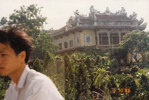 Bức ảnh chụp một ngôi chùa lớn theo kiến trúc cổ truyền Việt Nam, có thể nằm ở khu vực miền Trung như Huế hoặc Đà Nẵng, vào ngày 10 tháng 5 năm 1996.。**Thông tin văn bản:**。Trên các cột và dưới mái hiên của ngôi chùa có các ký tự, có thể là chữ Hán hoặc chữ Nôm, nhưng không thể đọc rõ do chất lượng ảnh. Ở góc dưới bên phải của ảnh có dấu ngày kỹ thuật số màu cam ghi '5 10 '96'.。**Mô tả cảnh quan và hoạt động:**。- **Loại địa điểm:** Đây là một ngôi chùa Phật giáo, tọa lạc trên một vị trí cao như đồi hoặc núi, được bao quanh bởi vườn cây.。- **Vật thể:**。- **Kiến trúc:** Một ngôi chùa nhiều tầng với tường sơn màu vàng. Mái nhà lợp ngói âm dương màu đỏ, có nhiều tầng và các góc mái cong vút. Trên nóc mái được trang trí rất công phu với các bức tượng làm bằng sành sứ hoặc điêu khắc, bao gồm hình rồng, phượng và bánh xe pháp luân, là biểu tượng đặc trưng của Phật giáo.。- **Thiên nhiên:** Ngôi chùa được bao quanh bởi cây cối xanh tươi, tạo thành một khu vườn. Có thể nhận ra một số cây chuối và nhiều loại cây cảnh, cây bụi khác.。- **Con người:** Ở phía tiền cảnh bên trái, một phần hình ảnh của một người đàn ông trẻ tuổi mặc áo sơ mi trắng xuất hiện trong khung hình.。- **Hoạt động:** Bức ảnh ghi lại một khoảnh khắc tham quan, ngắm cảnh tại một địa điểm tôn giáo. Không có hoạt động nghi lễ cụ thể nào đang diễn ra.。（日本語訳）。1996年5月10日に、ベトナムの伝統建築様式で建てられた大きなお寺を撮影した写真。フエやダナンなどの中部地域にある可能性があります。**テキスト情報：**。写真の右下隅には、オレンジ色のデジタル日付スタンプで「'96 5 10」と記されています。**風景と活動の説明：**。- **場所の種類：** これは仏教寺院であり、丘や山のような高台に位置し、庭園に囲まれています。- **物体：**。- **建築：** 黄色い壁を持つ多層構造の寺。屋根は赤色の陰陽瓦で葺かれており、多層で軒の角は反り上がっています。屋根の上には、龍、鳳凰、そして仏教の典型的な象徴である法輪など、陶磁器や彫刻で作られた像で非常に精巧に装飾されています。- **自然：** 寺は青々とした木々に囲まれ、庭園を形成しています。数本のバナナの木や、他の多くの観葉植物、低木が見て取れます。- **人物：** 左側の前景に、白いシャツを着た若い男性の姿の一部が写り込んでいます。- **活動：** この写真は、宗教的な場所での観光や景色を眺めている瞬間を記録したものです。特定の儀式活動は行われていません。