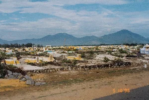 Bức ảnh chụp một khu nghĩa trang rộng lớn vào ngày 10 tháng 5 năm 1996. Khu nghĩa trang nằm trên một vùng đất cát, với rất nhiều ngôi mộ được xây dựng san sát nhau. Các ngôi mộ có nhiều kiểu dáng, kích thước và màu sắc đa dạng, chủ yếu là màu vàng, trắng và xanh lam. Một số ngôi mộ có bia đá nhưng không thể đọc rõ chữ khắc trên đó. Cây bụi mọc thưa thớt xen kẽ giữa các ngôi mộ. Ở phía sau khu nghĩa trang là dãy núi trập trùng dưới bầu trời có mây. Ở tiền cảnh là một bãi đất và mép của một con đường. Không có hoạt động nào của con người diễn ra, khung cảnh rất tĩnh lặng. Ở góc dưới cùng bên phải có dấu ngày tháng kỹ thuật số '5 10 '96'.。（日本語訳）。1996年5月10日に撮影された、広大な墓地の写真。墓地は砂地にあり、たくさんの墓が密集して建てられている。墓は様々な形、大きさ、色をしており、主に黄色、白、青である。いくつかの墓には墓石があるが、そこに刻まれた文字をはっきりと読み取ることはできない。墓の間には低木がまばらに生えている。墓地の後方には、曇り空の下に連なる山々がある。前景には空き地と道路の端がある。人の活動は見られず、景色は非常に静かである。右下隅に「'5 10 '96」というデジタル日付のスタンプがある。