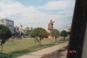 Bức ảnh chụp một cảnh quan đô thị ở Việt Nam vào ngày 10 tháng 5 năm 1996, có thể là một bùng binh hoặc một công viên nhỏ ven đường. Ở trung tâm của bãi cỏ xanh là một tượng đài lớn bằng đá, tạc hình một người đàn ông mặc áo choàng, đang giơ tay chỉ về phía trước. Xung quanh tượng đài và dọc theo bãi cỏ là những cây xanh nhỏ được cắt tỉa cẩn thận. Phía sau là một con đường có nhiều người dân đang di chuyển bằng xe đạp và xe máy. Dọc theo con đường là các tòa nhà nhiều tầng. Trên một tòa nhà màu xám, có một tấm biển quảng cáo lớn, có thể đọc được chữ "CÔNG TY", nhưng các dòng chữ còn lại bị mờ và không thể đọc rõ. Có thêm các biển hiệu khác trên các tòa nhà nhưng cũng không thể nhận dạng được nội dung. Bầu trời trong xanh và có mây. Bức ảnh dường như được chụp từ bên trong một phương tiện đang di chuyển.。（日本語訳）。1996年5月10日にベトナムで撮影された都市景観の写真で、ロータリーまたは道端の小さな公園のようです。緑の芝生の中央には、マントを羽織り、前方を指差している男性が彫られた大きな石像があります。像の周りと芝生に沿って、丁寧に手入れされた小さな植木があります。後方には道があり、多くの人々が自転車やバイクで移動しています。道沿いには複数階建ての建物が並んでいます。灰色の建物の一つには大きな広告看板があり、「CÔNG TY」という文字は読み取れますが、残りの文字はぼやけていてはっきりと読むことはできません。建物には他にも看板がありますが、その内容も識別できません。空は青く澄んでおり、雲があります。この写真は、移動中の乗り物の中から撮影されたようです。