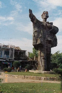 Bức ảnh chụp một tượng đài lớn bằng đá sẫm màu, đặt tại một khu vực công cộng như bùng binh hoặc công viên ở một đô thị. Tượng đài khắc họa một nhân vật chính, dường như là một người phụ nữ, trong tư thế hiên ngang với một cánh tay giơ lên. Trên thân tượng có chạm nổi chân dung của các nhân vật khác, bao gồm một người lính đội mũ. Tượng đài được đặt trên một bệ đá, xung quanh là bãi cỏ và lối đi. Phía sau là khung cảnh đường phố với những ngôi nhà nhiều tầng, trên mái nhà có các ăng-ten TV. Hoạt động của con người trong ảnh bao gồm một người đang lái xe máy, một số người đang ngồi nghỉ và có vài chiếc xe đạp dựng gần đó. Trên một tòa nhà có tấm biển hiệu ghi "HIỆU BUÔN ... Tài", nhưng một phần chữ đã bị che. Trên bệ tượng đài có một tấm bảng ghi chữ nhưng không thể đọc được nội dung. Góc dưới bên trái ảnh có dấu ngày tháng màu vàng là '96. 05. 10'.。（日本語訳）。写真は、ある都市のロータリーや公園のような公共エリアに設置された、暗い色の石でできた大きな記念碑を写している。記念碑は、片腕を上げた堂々とした姿の、女性と思われる一人の主要な人物を描写している。像の本体には、帽子をかぶった兵士を含む他の人物たちの肖像が浮き彫りにされている。記念碑は石の台座の上に設置されており、周囲には芝生と通路がある。後方には複数階建ての家々がある街の風景があり、屋根の上にはテレビアンテナがある。写真の中の人々の活動には、バイクを運転している一人、座って休憩している数人、そして近くに停められている数台の自転車が含まれる。ある建物には「HIỆU BUÔN ... Tài」と書かれた看板があるが、文字の一部は隠れている。記念碑の台座には文字が書かれたプレートがあるが、その内容は読み取れない。写真の左下隅には、「'96. 05. 10'」という黄色い日付の印がある。