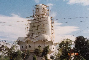 Bức ảnh ghi lại cảnh một công trình xây dựng một bức tượng Phật lớn ngoài trời. Bức tượng Phật màu trắng, trong tư thế ngồi thiền trên một đài sen lớn. Toàn bộ phần thân trên và đầu của bức tượng được bao quanh bởi một hệ thống giàn giáo bằng kim loại, cho thấy công trình đang trong giai đoạn thi công hoặc trùng tu. Phần lớn bức tượng được che phủ bởi một lớp vải hoặc bạt màu trắng. Đài sen ở phía dưới có các ô cửa sổ nhỏ hình tròn. Xung quanh chân tượng có nhiều cây cối xanh tươi. Phía sau là bầu trời xanh có mây. Một vài đường dây điện chạy ngang qua khung hình. Ở góc dưới bên phải của hình ảnh có dấu ngày tháng màu cam ghi '5 10 '96', cho thấy bức ảnh được chụp vào ngày 10 tháng 5 năm 1996.。（日本語訳）。写真は、屋外で大きな仏像を建設している工事現場の光景を記録しています。仏像は白色で、大きな蓮華座の上で座禅を組む姿勢です。像の上半身と頭部は全体が金属製の足場システムで囲まれており、工事が建設または修復の段階にあることを示しています。像の大部分は白い布またはシートで覆われています。下部の蓮華座には小さな円形の窓があります。像の足元周辺には多くの青々とした木々があります。後ろには雲のある青空が広がっています。数本の電線がフレームを横切っています。画像の右下隅にはオレンジ色の日付スタンプ「'5 10 '96」があり、写真が1996年5月10日に撮影されたことを示しています。