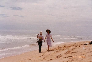 Bức ảnh chụp cảnh hai người phụ nữ đang đi dạo trên một bãi biển vào một ngày trời nhiều mây. Bối cảnh là một bờ biển rộng lớn với bãi cát vàng và những con sóng đang xô vào bờ.。**Chi tiết cảnh vật và hoạt động:**。*   **Địa điểm:** Một bãi biển, có khả năng là ở khu vực miền Trung Việt Nam như Huế hoặc Đà Nẵng.。*   **Hoạt động:** Hai người phụ nữ đang đi dạo dọc theo mép nước trên bãi cát. Người phụ nữ bên trái cầm một chiếc máy ảnh, cho thấy họ có thể đang đi du lịch và chụp ảnh kỷ niệm. Không khí trong ảnh mang lại cảm giác thư thái, yên bình.。*   **Con người và vật thể:**。*   Hai người phụ nữ. Người phụ nữ bên trái mặc áo phông trắng có họa tiết, quần dài tối màu, đội mũ tai bèo màu trắng, và đeo một chiếc túi nhỏ.。*   Người phụ nữ bên phải mặc một chiếc váy trắng dài đến bắp chân, tay phồng và đội một chiếc mũ rộng vành màu sẫm.。*   Phong cảnh bao gồm biển cả với sóng bạc đầu, bãi cát có nhiều dấu chân, và bầu trời u ám.。*   Ở góc xa bên phải, có thể thấy một vài vật dụng cá nhân và rác như túi ni lông trên cát.。**Thông tin chữ viết:**。*   Ở góc dưới cùng bên phải của bức ảnh có một dấu ngày tháng kỹ thuật số màu vàng, ghi là "11 9 '96".。（日本語訳）。曇りの日にビーチを散歩している二人の女性の風景を撮った写真です。背景は、黄色い砂浜と岸に打ち寄せる波がある広大な海岸です。**風景と活動の詳細：**。*   **場所：** ビーチ。フエやダナンなど、ベトナム中部の地域である可能性があります。*   **活動：** 二人の女性が砂浜の水際に沿って散歩しています。左側の女性はカメラを持っており、彼女たちが旅行中で記念写真を撮っている可能性を示しています。写真の雰囲気は、リラックスした穏やかな感覚をもたらします。*   **人物と物体：**。*   二人の女性。左側の女性は、柄の入った白いTシャツ、暗い色の長ズボンを着用し、白いバケットハットをかぶり、小さなバッグを身に着けています。*   右側の女性は、ふくらはぎ丈の白いワンピースを着ており、袖はパフスリーブで、暗い色のつばの広い帽子をかぶっています。*   風景には、白波の立つ海、多くの足跡がある砂浜、そして曇り空が含まれています。*   右奥の隅には、砂の上にビニール袋のような個人の持ち物やゴミがいくつか見えます。**文字情報：**。*   写真の右下隅には、「11 9 '96」と記された黄色いデジタルの日付スタンプがあります。