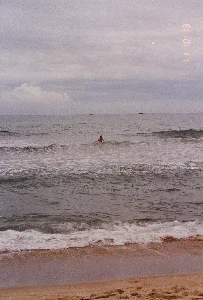 Dấu thời gian được in trên ảnh là '11 10:13'. Bức ảnh chụp cảnh một bãi biển vào một ngày trời nhiều mây. Ở phía trước là bãi cát, với những con sóng đang vỗ vào bờ. Giữa biển, một người đàn ông đang tắm. Xa xa trên đường chân trời, có thể nhìn thấy bóng dáng của một vài chiếc thuyền nhỏ, có khả năng là thuyền đánh cá.。（日本語訳）。写真に印刷されているタイムスタンプは「11 10:13」です。写真は曇りの日のビーチの光景を撮影したものです。手前には砂浜があり、波が岸に打ち寄せています。海の中では、一人の男性が海水浴をしています。遠くの水平線上には、数隻の小さな船の影が見え、おそらく漁船だと思われます。