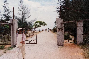 Bức ảnh chụp tại một lối vào có cổng lớn dẫn ra một con đường đất. Ở phía trước, một người phụ nữ đội nón lá, mặc quần áo sáng màu và áo khoác mỏng, đang đi bộ ra khỏi cổng. Cổng được xây bằng hai trụ đá hoa cương và cửa sắt đang mở. Trên trụ cổng bên phải có một tấm biển nhưng chữ viết đã bị mờ, không thể đọc được. Xung quanh có nhiều cây cối, đặc biệt là cây phi lao, cho thấy đây có thể là một khu vực ven biển. Ở phía sau, trên con đường đất, có một vài người đang đi lại, một chiếc xe máy, một chiếc xe đạp và một chiếc ô tô đang đậu ở phía xa, hướng ra biển. Bầu trời có mây. Ở góc dưới bên phải của bức ảnh có dấu thời gian '11 11:00'.。（日本語訳）。写真は、未舗装の道に通じる大きな門のある入り口で撮影されたものです。手前では、ノンラーをかぶり、明るい色の服と薄い上着を着た女性が、門から歩いて出てきています。門は2本の大理石の柱と鉄の扉でできており、開いています。右側の門柱には看板がありますが、文字がかすれていて読むことができません。周囲には多くの木々、特にモクマオウがあり、ここが沿岸地域である可能性を示しています。奥の未舗装の道には、数人が行き交い、バイク1台、自転車1台、そして遠くに海の方を向いて駐車している車が1台あります。空は曇っています。写真の右下隅には「11 11:00」というタイムスタンプがあります。