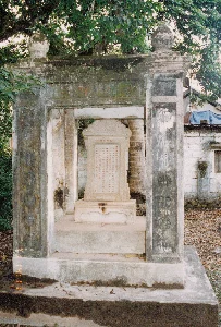 Bức ảnh chụp một công trình kiến trúc cổ ngoài trời, dường như là một nhà bia tưởng niệm hoặc một phần của một khu lăng mộ. Công trình được xây bằng đá và xi măng, đã cũ kỹ và nhuốm màu thời gian với rêu phong bám trên bề mặt. Cấu trúc chính bao gồm một tấm bia đá đặt ở trung tâm, được che chắn bởi một kiến trúc có bốn cột và mái che. Tấm bia đá được đặt trên một bệ đá, trên đó khắc dày đặc các ký tự chữ Hán theo hàng dọc. Do chất lượng ảnh và sự bào mòn, nội dung chi tiết của văn bia không thể đọc được, nhưng có thể nhận ra đây là một bia ký ghi lại công trạng hoặc tiểu sử. Các cột trụ của nhà bia cũng có các họa tiết điêu khắc, có thể là các chữ Hán cách điệu, nhưng cũng đã bị mờ đi rất nhiều. Bối cảnh xung quanh là cây cối xanh tốt, có một thân cây (giống cây cau hoặc dừa) nằm ngay sau tấm bia. Ở phía sau bên phải, có một ngôi nhà tường trắng, mái nhà được che một phần bằng tấm bạt màu xanh. Mặt đất xung quanh công trình phủ đầy lá cây khô. Không có người hay hoạt động nào được ghi lại trong ảnh, tạo nên một không khí tĩnh lặng và trang nghiêm.。（日本語訳）。この建造物は石とセメントで建てられており、古びており、表面には苔が生え、時の流れを感じさせる。主な構造は中央に置かれた一枚の石碑を含み、4本の柱と屋根を持つ建築物によって覆われている。石碑は石の台座の上に置かれ、その上には縦書きの漢字がびっしりと刻まれている。写真の画質と摩耗により、碑文の詳細な内容は読み取れないが、これが功績や伝記を記録した碑文であることは認識できる。碑堂の柱にも彫刻模様があり、それはデザイン化された漢字かもしれないが、それもまた非常に薄れてしまっている。周囲の背景には青々とした木々があり、石碑のすぐ後ろには一本の木（ビンロウジュかヤシの木に似ている）の幹がある。右奥には、白い壁の家があり、その屋根は青いシートで一部が覆われている。建造物の周りの地面は枯れ葉で覆われている。写真には人や活動は記録されておらず、静かで荘厳な雰囲気を醸し出している。