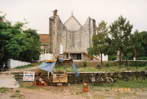 Bức ảnh chụp quang cảnh phía trước một nhà thờ lớn bằng đá, có kiến trúc hiện đại, đây là Nhà thờ Phủ Cam ở Huế. Nhà thờ có mặt tiền độc đáo với hai phần mái vươn lên cao và cong. Có một bức tượng Đức Mẹ Maria màu trắng đặt ở phía trước lối vào và một tượng nhỏ hơn trên đỉnh mái.。Ở tiền cảnh, có một quán nước nhỏ, tạm bợ được dựng dưới một chiếc ô lớn và những tấm bạt màu xanh. Trên một kệ gỗ, nhiều loại nước giải khát đóng chai và đồ ăn vặt được bày bán. Một nhóm người, bao gồm phụ nữ và trẻ em, đang ngồi trên những chiếc ghế nhựa đỏ thấp, có vẻ như đang nghỉ ngơi và uống nước. Bên cạnh quán có một tấm biển quảng cáo dịch vụ chụp ảnh, trên đó có ghi:。"PHOTO CAMERA。Duy Kỳ。Nhận CHỤP TIỆC HỘI。CƯỚI SINH NHẬT HỘI NGHỊ。Ép Plastic"。Trong khung cảnh còn có các phương tiện đặc trưng của thập niên 90 ở Việt Nam: một chiếc xe máy đỗ ở bên trái và một chiếc xe ba bánh (giống xe xích lô) ở bên phải. Xung quanh là bãi đất và cỏ, với nhiều cây xanh như tre và thông. Khung cảnh phản ánh một nét sinh hoạt đời thường, bình dị tại một địa danh tôn giáo nổi tiếng vào năm 1996.。（日本語訳）。写真は、現代的な建築様式の石造りの大きな教会の前景を写したもので、これはフエのフーカム教会です。教会は、2つの屋根部分が高く伸びて湾曲したユニークなファサードを持っています。入り口の前には白い聖母マリア像が置かれ、屋根の頂上にはより小さな像があります。前景には、大きな傘と青い防水シートの下に建てられた、小さく簡素な水飲み屋があります。木製の棚には、多くの種類のボトル入り飲料やスナック菓子が並べられて売られています。女性や子供を含む一団の人々が、低い赤いプラスチックの椅子に座っており、休憩して水を飲んでいるように見えます。店の隣には写真撮影サービスを宣伝する看板があり、そこには次のように書かれています：。「フォトカメラ。ズイ・キー。パーティー・宴会の撮影を承ります。結婚式 誕生日 会議。ラミネート加工」。風景の中には、90年代のベトナムに特徴的な乗り物もあります。左側には一台のバイクが駐車しており、右側には一台の三輪車（シクロに似た）があります。周囲は土と草の広場で、竹や松のような多くの緑の木々があります。この光景は、1996年における有名な宗教的名所での、日常的で素朴な生活の一面を反映しています。