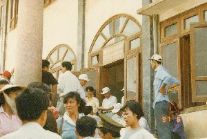 Bức ảnh chụp một đám đông người đang tụ tập trước cửa ra vào của một tòa nhà vào ngày 14 tháng 6 năm 1997. Dựa vào tấm biển hiệu, đây là một trụ sở hải quan tại một cửa khẩu biên giới. Phía trên cửa ra vào có một tấm biển ghi bằng ba thứ tiếng: "NƠI LÀM THỦ TỤC HẢI QUAN" (tiếng Việt), "PLACE OF GOING THROUGH CUSTOMS" (tiếng Anh, chữ hơi mờ), và "办理海关手续处" (tiếng Trung). Hoạt động chính đang diễn ra là mọi người chờ đợi để làm thủ tục xuất nhập cảnh hoặc thông quan. Trong đám đông có cả nam và nữ với trang phục đa dạng. Có một người phụ nữ đội nón lá và dùng khăn che mặt, một số người đàn ông đội mũ lưỡi trai, và một người đội mũ kepi màu xanh lá cây, có thể là sĩ quan hoặc bộ đội. Tòa nhà có kiến trúc với các cột trụ, cửa sổ và cửa ra vào bằng gỗ, phần trên cửa sổ có dạng vòm.。（日本語訳）。1997年6月14日、ある建物の出入り口の前に集まっている人々の群衆を写した写真です。看板から、これは国境検問所の税関であることがわかります。出入り口の上には、「NƠI LÀM THỦ TỤC HẢI QUAN」（ベトナム語）、「PLACE OF GOING THROUGH CUSTOMS」（英語、文字は少しぼやけている）、そして「办理海关手续处」（中国語）と3つの言語で書かれた看板があります。主に行われている活動は、人々が出入国手続きまたは税関手続きを待っていることです。群衆の中には、様々な服装をした男性と女性がいます。ノンラーをかぶりスカーフで顔を覆っている女性が一人、野球帽をかぶっている男性が数人、そして緑色のケピ帽をかぶっている人が一人おり、将校か兵士である可能性があります。建物は柱、木製の窓と出入り口があり、窓の上部はアーチ状になっている建築様式です。