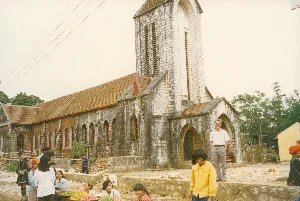 Bối cảnh là khu vực phía trước Nhà thờ đá Sa Pa ở tỉnh Lào Cai, được chụp vào ngày 14 tháng 6 năm 1997. Đây là một công trình kiến trúc cổ bằng đá theo phong cách Gothic với tháp chuông, mái lợp ngói và các cửa sổ hình vòm. Các bức tường đá đã nhuốm màu thời gian. Một phiên chợ nhỏ đang diễn ra trên khoảng sân đất phía trước nhà thờ. Một số phụ nữ và trẻ em, trong đó có người mặc trang phục dân tộc thiểu số đặc trưng của vùng Sa Pa, đang ngồi bán các loại trái cây (có thể là mận hoặc đào) được bày trong những chiếc rổ. Một người đàn ông nước ngoài đang đứng tạo dáng chụp ảnh. Nhiều người khác đang tụ tập và đi lại trong khu vực. Phía trên có các đường dây điện. Không có văn bản nào có thể đọc được từ các biển hiệu hay bảng chỉ dẫn, nhưng có dấu ngày tháng trên ảnh là '97 6 14'.。（日本語訳）。背景は、1997年6月14日に撮影された、ラオカイ省サパの石造教会前のエリアです。これは、鐘楼、瓦葺きの屋根、アーチ型の窓を持つ、ゴシック様式の古い石造りの建築物です。石の壁は時の色を帯びています。教会の前の土の広場で小さな市が開かれています。サパ地方特有の少数民族の衣装を着た人々を含む数人の女性と子供たちが、籠に盛られた果物（おそらくプラムか桃）を座って売っています。一人の外国人男性が写真撮影のためにポーズをとって立っています。他の多くの人々がそのエリアに集まったり行き来したりしています。上には電線があります。看板や案内板から読み取れる文字はありませんが、写真には「'97 6 14」という日付のスタンプがあります。