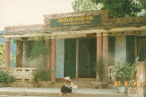 Bức ảnh chụp mặt tiền của một tòa nhà một tầng, là Trạm Kiểm soát Đồn Biên phòng Cửa khẩu Lào Cai, vào ngày 14 tháng 6 năm 1997. Trên mái hiên có một tấm biển lớn màu xanh lá cây với dòng chữ màu vàng: 'TRẠM KIỂM SOÁT ĐỒN BIÊN PHÒNG CỬA KHẨU LÀO CAI'. Phía trên một lối vào, có một tấm biển nhỏ hơn màu đỏ ghi 'P. TIẾP KHÁCH', có thể là viết tắt của 'Phòng Tiếp Khách'. Tòa nhà có kiến trúc kiểu cũ với hàng hiên rộng và các cột trụ tròn sơn màu đỏ gạch. Các bức tường đã cũ và bạc màu, cửa ra vào và cửa sổ là loại cửa chớp gỗ màu xanh lá. Ở phía trước, một người phụ nữ đội nón lá, mặc trang phục đơn giản, đang ngồi nghỉ trên bậc thềm. Bên cạnh bà có một chiếc túi ni lông. Xung quanh tòa nhà có nhiều cây cối. Trên hiên có đặt vài chậu cây cảnh, trong đó có một chậu sứ men xanh trắng. Khung cảnh cho thấy một không gian yên tĩnh tại một cơ quan ở khu vực biên giới.。（日本語訳）。1997年6月14日に撮影された、ラオカイ国境ゲート国境警備隊駐屯地検問所である、1階建ての建物の正面を写した写真。軒先には、「TRẠM KIỂM SOÁT ĐỒN BIÊN PHÒNG CỬA KHẨU LÀO CAI」と黄色い文字で書かれた大きな緑色の看板がある。ある入口の上には、「P. TIẾP KHÁCH」と書かれた小さな赤い看板があり、これは「Phòng Tiếp Khách」（応接室）の略である可能性がある。建物は古い様式の建築で、広いベランダとレンガ色に塗られた丸い柱がある。壁は古く色褪せており、出入口のドアと窓は緑色の木製よろい戸タイプである。正面では、ノンラーをかぶり、簡素な服装をした一人の女性が、階段に座って休んでいる。彼女の隣にはビニール袋が一つある。建物の周りには多くの木々がある。ベランダにはいくつかの観葉植物の鉢が置かれており、その中には青と白の磁器の鉢が一つある。この光景は、国境地域の官庁における静かな空間を示している。