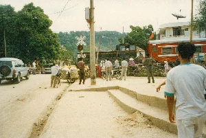 Bối cảnh là một điểm giao cắt đường sắt ở một khu vực có vẻ là thị trấn hoặc vùng ven đô tại Việt Nam, vào khoảng năm 1997. Một đoàn tàu hỏa đang chắn ngang đường, khiến người dân và các phương tiện phải dừng lại chờ đợi. Con đường không được trải nhựa, là đường đất, và có một vài bậc thềm bê tông dẫn lên vỉa hè.。Các dòng chữ có thể nhận diện được bao gồm:。- Trên biển báo giao thông đường sắt hình chữ thập (dấu St. Andrew): "CHÚ Ý TÀU LỬA".。- Trên biển báo phụ hình chữ nhật bên dưới: "ĐÈN ĐỎ DỪNG LẠI".。- Trên toa tàu màu đỏ: "T.B - 21761".。- Trên đầu máy hoặc toa tàu màu sẫm phía trước: có thể đọc được một phần là "D4H-411".。Hoạt động chính là mọi người đang chờ tàu đi qua. Có nhiều người đi xe máy, xe đạp và người đi bộ. Một số phụ nữ đội nón lá. Nổi bật có một người đàn ông đang đẩy một chiếc xe đạp thồ, chở theo hai chiếc sọt tre/mây rất lớn chứa đầy hàng hóa và được phủ vải trắng. Ở phía bên trái, một chiếc ô tô địa hình màu bạc đang đỗ. Phía xa, một tòa nhà có treo cờ Việt Nam và được lắp đặt một chảo thu sóng vệ tinh.。（日本語訳）。背景は1997年頃のベトナムで、町か郊外と思われる地域にある鉄道の踏切です。一本の列車が道路を横切って塞いでおり、人々や乗り物は停止して待たなければなりません。道路は舗装されておらず、土の道で、歩道へと続くいくつかのコンクリートの段差があります。認識できる文字には以下が含まれます：。- 十字形（聖アンデレ十字）の鉄道交通標識上：「CHÚ Ý TÀU LỬA」。- その下の長方形の補助標識上：「ĐÈN ĐỎ DỪNG LẠI」。- 赤い車両上：「T.B - 21761」。- 前方の暗い色の機関車または車両上：部分的に「D4H-411」と読み取れます。主な活動は、人々が列車が通り過ぎるのを待っていることです。バイクや自転車に乗る人、そして歩行者がたくさんいます。何人かの女性はノンラーをかぶっています。目立っているのは、荷物運搬用の自転車を押している一人の男性で、品物でいっぱいで白い布で覆われた、非常に大きな竹か籐で編んだ籠を二つ運んでいます。左側には、一台の銀色のオフロード車が駐車しています。遠くには、ベトナム国旗が掲げられ、衛星放送受信用のパラボラアンテナが設置された建物があります。