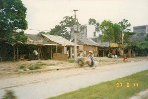 Bức ảnh chụp cảnh sinh hoạt đời thường ven một con đường đất tại một vùng quê hoặc thị trấn nhỏ ở Việt Nam, được ghi lại vào ngày 14 tháng 6 năm 1997.。**Thông tin chữ viết:**。- Trên một ngôi nhà bên trái có treo tấm biển màu đỏ với chữ trắng, có thể nhận dạng được chữ "CẮT TÓC".。- Ở phía xa hơn có một biển hiệu dựng dọc màu xanh và vàng, nhưng chữ viết trên đó không thể đọc được do bị mờ.。- Dấu ngày tháng màu vàng ở góc dưới bên phải của bức ảnh ghi rõ "97 6 14".。**Mô tả cảnh vật và hoạt động:**。- **Địa điểm:** Khung cảnh diễn ra trên một con đường đất, chưa được trải nhựa. Dọc bên đường là dãy nhà cấp 4 liền kề với kiến trúc đơn sơ, mái lợp tôn. Những ngôi nhà này có vẻ vừa là nhà ở, vừa là các cửa hàng nhỏ kinh doanh ven đường.。- **Hoạt động:** Một người đội nón lá đang đạp một chiếc xe đạp trên đường. Phía sau xe chở nhiều hàng hóa, cho thấy hoạt động vận chuyển hàng hóa bằng phương tiện thô sơ. Một người khác đang đứng cạnh một chiếc xe đạp khác. Dọc ven đường, một vài người dân địa phương, bao gồm cả trẻ em, đang đứng, ngồi nghỉ ngơi hoặc sinh hoạt trước hiên nhà. Không khí chung cho thấy một buổi chiều yên bình ở vùng nông thôn.。- **Vật thể:** Các vật thể nổi bật bao gồm xe đạp, nón lá, những ngôi nhà cấp 4, và một cột điện bê tông lớn có gắn máy biến áp ở trung tâm, đặc trưng cho cơ sở hạ tầng của thời kỳ đó.。（日本語訳）。1997年6月14日に記録された、ベトナムの田舎または小さな町の未舗装の道端での日常生活の風景を撮影した写真。**文字情報：**。- 左側の家には、白い文字で「CẮT TÓC」（散髪）と識別できる赤い看板が掲げられています。- さらに奥には青と黄色の縦長の看板がありますが、その上の文字はぼやけていて読むことができません。- 写真の右下隅にある黄色の日付スタンプには、「97 6 14」と明確に記されています。**風景と活動の説明：**。- **場所：** 風景は、まだ舗装されていない土の道で展開されています。道沿いには、簡素な建築様式でトタン屋根の平屋が隣接して並んでいます。これらの家々は、住居であると同時に、道端で商売をする小さな店のようです。- **活動：** ノンラー（ベトナムの伝統的な笠）をかぶった人が道で自転車を漕いでいます。自転車の後ろには多くの荷物が積まれており、簡素な手段による物資輸送の様子がうかがえます。別の人がもう一台の自転車の隣に立っています。道端沿いでは、子供を含む数人の地元住民が、家の軒先で立ったり、座って休んだり、あるいは生活したりしています。全体的な雰囲気は、田舎の穏やかな午後を示しています。- **物体：** 目立つ物体には、自転車、ノンラー、平屋建ての家々、そして中央にある変圧器が取り付けられた大きなコンクリートの電柱が含まれ、これらはその時代のインフラを特徴づけています。