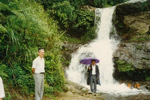 Bức ảnh ghi lại một khung cảnh thiên nhiên tại một thác nước ở vùng núi, có khả năng là khu vực Sa Pa. Phong cảnh được bao phủ bởi cây cối xanh tươi và rậm rạp, bao gồm cả dương xỉ và các loại cây bụi khác. Dòng thác trắng xóa đang đổ mạnh xuống những tảng đá lớn, ẩm ướt.。Trong ảnh có hai người đàn ông đang tạo dáng chụp ảnh. Bên trái, một người đàn ông lớn tuổi, có thể là người nước ngoài, mặc áo sơ mi trắng cộc tay và quần tây màu xám. Ông đội một chiếc băng đô có hoa văn trên đầu và đang mỉm cười. Ở trung tâm, một người đàn ông khác đang đứng trên đá gần chân thác, cầm một chiếc ô (dù) lớn màu tím. Ông mặc áo khoác dài tối màu bên ngoài áo sơ mi trắng.。Ở góc dưới cùng bên phải của bức ảnh có các chữ số màu cam "6 15", đây có thể là dấu ngày tháng (ngày 15 tháng 6) do máy ảnh tự động in lên.。（日本語訳）。この写真は、山岳地帯にある滝の自然な風景を捉えており、おそらくサパ地域のものでしょう。風景は、シダやその他の種類の低木を含む、青々として鬱蒼とした木々で覆われています。白い滝が、大きく湿った岩々に力強く流れ落ちています。写真には、ポーズをとって写真を撮っている2人の男性がいます。左側には、年配の男性がおり、おそらく外国人でしょう。彼は白い半袖シャツと灰色のズボンを着用しています。彼は頭に模様の入ったヘッドバンドを巻いており、微笑んでいます。中央では、別の男性が滝の麓近くの岩の上に立っており、大きな紫色の傘を差しています。彼は白いシャツの上に暗い色の長いコートを着ています。写真の右下隅にはオレンジ色の数字「6 15」があり、これはカメラによって自動的に印字された日付（6月15日）の可能性があります。