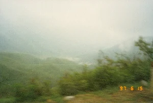 Có dòng chữ số màu vàng ở góc dưới bên phải ghi ngày tháng: "97 6 15", tức là ngày 15 tháng 6 năm 1997. Bức ảnh được chụp từ một phương tiện đang di chuyển, ghi lại cảnh quan một vùng núi cao. Toàn bộ khung cảnh chìm trong sương mù dày đặc. Tiền cảnh là các bụi cây ven đường bị nhòe do chuyển động. Hậu cảnh là những dãy núi trập trùng, mờ ảo trong sương. Ở phía xa, có thể thấy một thung lũng hoặc một khu vực sáng hơn nhưng không rõ chi tiết.。（日本語訳）。右下隅に「97 6 15」という黄色い数字の列があり、1997年6月15日を意味しています。写真は移動中の乗り物から撮影されたもので、高山地帯の風景を記録しています。風景全体が濃い霧に包まれています。前景には、動きによってぼやけた道端の茂みがあります。遠くには、谷またはより明るい領域が見えますが、詳細は不明瞭です。