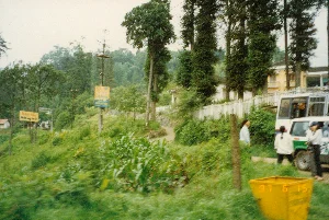 Bối cảnh là một con đường ở vùng núi, có thể là ở khu vực Sa Pa, Lào Cai, vào năm 1997. Phong cảnh bao gồm đồi núi trập trùng với cây cối xanh tươi và thảm thực vật rậm rạp, bao gồm cả một đám cây ngô ở phía trước.。Về chữ viết, có hai biển báo ven đường nhưng chữ viết rất mờ. Một biển báo màu vàng bên trái có vẻ ghi 'Điểm Tạm', các chữ còn lại không đọc được. Biển báo còn lại ở giữa, có phần trên màu vàng và phần dưới màu xanh, cũng không thể đọc rõ. Dưới góc phải bức ảnh, một thùng rác màu vàng có dấu ngày tháng kỹ thuật số '97 6 15'.。Về cảnh vật và hoạt động, một con đường mòn đất nhỏ dẫn lên dốc, bên cạnh là hàng rào cọc màu trắng. Phía sau hàng rào là một ngôi nhà hai tầng sơn vàng. Bên phải, một chiếc xe khách nhỏ màu trắng và xanh lá cây đang đỗ. Có ba người đang đứng gần chiếc xe, có vẻ đang chờ đợi hoặc trò chuyện. Một người khác đang đi bộ trên con đường mòn. Khung cảnh thể hiện một sinh hoạt đời thường ở vùng cao.。（日本語訳）。背景は山岳地帯の道で、おそらく1997年のラオカイ省サパ地区でしょう。風景には、緑豊かな木々と鬱蒼とした植生が広がる起伏のある丘や山々が含まれており、手前にはトウモロコシ畑もあります。文字については、道端に2つの看板がありますが、文字は非常にぼやけています。左側の黄色い看板には「Điểm Tạm（一時的な地点）」と書かれているようですが、残りの文字は読めません。中央にあるもう一つの看板は、上部が黄色で下部が青色ですが、これもはっきりと読むことはできません。写真の右下には、黄色いゴミ箱に「'97 6 15」というデジタル日付のスタンプがあります。風景と活動については、小さな土の道が坂を上っており、その隣には白い杭の柵があります。柵の後ろには、黄色く塗られた2階建ての家があります。右側には、白と緑の小さなバスが駐車しています。車の近くに3人が立っており、待っているか、おしゃべりをしているようです。もう一人が小道を歩いています。この光景は、高地での日常的な生活の一場面を表しています。