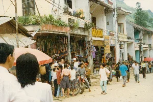 Bức ảnh chụp một con phố chợ nhộn nhịp ở một thị trấn miền núi, có thể là Sa Pa, vào khoảng năm 1997. Con đường không được trải nhựa mà vẫn là đường đất. Dọc hai bên đường là những dãy nhà phố 2-3 tầng, tầng trệt được dùng làm cửa hàng và nhà nghỉ.。Các dòng chữ trên các biển hiệu có thể nhận dạng được bao gồm:。- Biển hiệu màu xanh: "LOTUS HOTEL", "WELCOME".。- Biển hiệu màu vàng: "NHÀ NGHỈ THÁC BẠC", "Water Fall GUEST HOUSE", "WELCOME", và số điện thoại "TEL: 020..." (mã vùng cũ của Lào Cai).。Hoạt động chính là một phiên chợ. Rất đông người dân địa phương, bao gồm cả phụ nữ dân tộc thiểu số mặc trang phục truyền thống và gùi những chiếc giỏ tre lớn sau lưng, đang tụ tập mua bán và đi lại trên phố. Có cả một vài du khách nước ngoài trong đám đông. Các cửa hàng treo bán rất nhiều mặt hàng thổ cẩm như áo, váy, túi xách với hoa văn sặc sỡ. Một người đàn ông đang ngồi trên chiếc xe máy màu đỏ. Một số người đi bộ dùng ô (dù) màu đỏ để che nắng. Khung cảnh chung cho thấy một sinh hoạt đời thường sôi động và đặc trưng của một thị trấn du lịch vùng cao Việt Nam thời kỳ đó.。（日本語訳）。写真は、1997年頃、山岳地帯の町、おそらくサパの賑やかな市場の通りを写したものです。道路は舗装されておらず、まだ土の道です。道沿いの両側には2〜3階建ての商店が立ち並び、1階は店舗やゲストハウスとして使われています。認識できる看板の文字には以下が含まれます：。- 青い看板：「LOTUS HOTEL」、「WELCOME」。- 黄色い看板：「NHÀ NGHỈ THÁC BẠC」（タックバック・ゲストハウス）、「Water Fall GUEST HOUSE」、「WELCOME」、そして電話番号「TEL: 020...」（ラオカイの旧市外局番）。主な活動は市場です。伝統的な衣装を身に着け、背中に大きな竹籠を背負った少数民族の女性を含む多くの地元の人々が、通りに集まって売買したり行き来したりしています。人混みの中には数人の外国人観光客もいます。店先には、色鮮やかな模様のシャツ、スカート、バッグなどの錦織製品が多数吊るされて売られています。一人の男性が赤いバイクに座っています。何人かの歩行者は、日差しを避けるために赤い傘を使っています。全体的な光景は、その時代のベトナムの高原にある観光地の、活気に満ちた特徴的な日常生活を示しています。