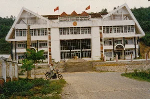 Bức ảnh chụp một tòa nhà hành chính lớn, nhiều tầng, sơn màu trắng với kiến trúc đối xứng đặc trưng, có hai mái hình tam giác lớn ở hai bên. Dựa vào bối cảnh địa lý (Lào Cai) và các biểu tượng, đây rất có thể là tòa nhà Cửa khẩu Quốc tế Lào Cai vào thời điểm năm 1997. Trên mặt tiền tòa nhà có gắn Quốc huy nước Cộng hòa Xã hội Chủ nghĩa Việt Nam. Trên nóc nhà có hai lá cờ đang bay, một là Cờ Tổ quốc (cờ đỏ sao vàng) và một là cờ Đảng Cộng sản Việt Nam (cờ búa liềm).。Phía trước tòa nhà là một con đường chưa được trải nhựa hoàn toàn. Một người đàn ông đội mũ đang điều khiển một chiếc xe máy kiểu cũ. Để đi vào tòa nhà, phải đi lên một cầu thang bộ rộng. Trên khu vực cầu thang có một vài người và một đống vật liệu xây dựng như sỏi đá, cho thấy công trình có thể mới được xây dựng hoặc đang được sửa chữa. Khung cảnh xung quanh là đồi núi và cây cối xanh tươi.。（日本語訳）。写真には、白く塗られた、特徴的な左右対称の建築様式を持つ、多層階の大きな行政庁舎が写っており、両側には大きな三角形の屋根が2つあります。地理的背景（ラオカイ）と象徴物から、これは1997年当時のラオカイ国際口岸の建物である可能性が非常に高いです。建物の正面には、ベトナム社会主義共和国の国章が取り付けられています。屋根の上には2つの旗がはためいており、1つは祖国旗（金星紅旗）、もう1つはベトナム共産党旗（鎌と槌の旗）です。建物の前には、まだ完全には舗装されていない道があります。帽子をかぶった男性が、旧式のバイクを運転しています。建物に入るには、広い階段を上らなければなりません。階段のエリアには数人の人と、砂利や石のような建設資材の山があり、この建物が新築されたばかりか、あるいは修繕中であることを示しています。周囲の風景は、丘や山、そして青々とした木々です。