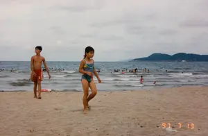 Góc dưới bên phải của ảnh có dấu ngày tháng màu cam: '97 7 13'.。Đây là khung cảnh tại một bãi biển vào một ngày trời nhiều mây. Ở phía trước, trên bãi cát, có hai đứa trẻ. Một bé trai cởi trần, mặc quần bơi màu đỏ có sọc trắng và chữ 'B', đang đi bộ trên cát. Bên cạnh là một bé gái mặc đồ bơi một mảnh nhiều màu sắc, tóc buộc sau, cũng đang đi bộ. Ở phía sau, dưới biển có rất nhiều người đang tắm và chơi đùa với sóng. Một số người đang sử dụng phao bơi. Xa xa trên mặt biển là hình ảnh những ngọn núi. Trên bãi cát, phía sau cậu bé, có một chiếc phao cứu sinh màu đỏ và trắng. Hoạt động chính đang diễn ra là mọi người đi nghỉ mát và tắm biển.。（日本語訳）。写真の右下隅に、オレンジ色の「'97 7 13」という日付スタンプがあります。これは曇りの日のビーチの光景です。手前の砂浜には、2人の子供がいます。男の子は上半身裸で、白いストライプと「B」の文字が入った赤い海水パンツを履いて、砂の上を歩いています。その隣には、カラフルなワンピース水着を着て髪を後ろで結んだ女の子がいて、彼女も歩いています。後方では、海の中にたくさんの人がいて、海水浴をしたり波と戯れたりしています。何人かは浮き輪を使っています。遠くの海面には、山々の姿が見えます。砂浜の、男の子の後ろには、赤と白の救命浮き輪があります。行われている主な活動は、人々が休暇を過ごし、海水浴をしていることです。